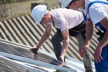 Male engineers installing stand-alone solar photovoltaic panel system. Two electricians mounting blue solar module on roof of modern house. Alternative energy environmental concept.