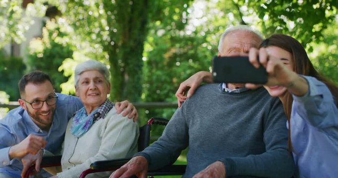 Slow motion of happy grandchildren and grandparents both in a wheelchair are having fun to take a selfie photo with cellular phone or making the video call  grandparents both in a wheelchair in a park