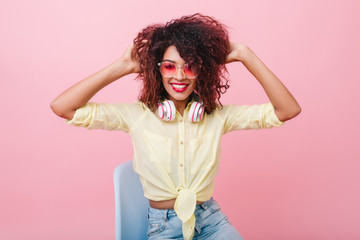 Terrific african girl in pink sunglasses sitting in blue chair. Laughing black female model in cotton shirt playing with curly brown hair.