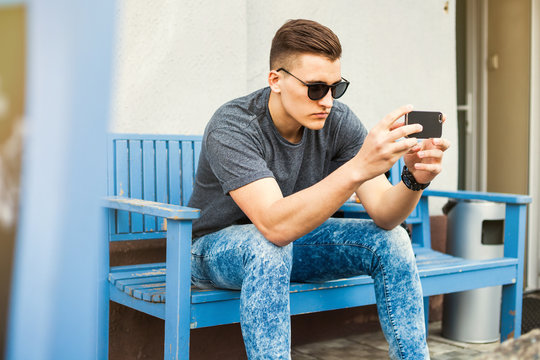 Hipster style guy with and a fashionable hairdo. a young man sitting on a bench in the street and make a photo, vintage photo.In the hands of a fashion backpack and a lion, sunglasses, sneakers, jeans