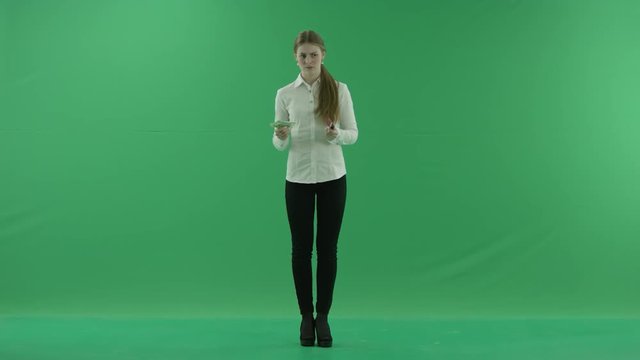 Young business woman is holding cash and credit card in each hand and choosing, when showing plastic card. She wears formal dress: white shirt and blak trousers and stands on the green screen.