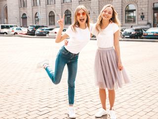 Portrait of two young beautiful blond smiling hipster girls in trendy summer white t-shirt clothes. Sexy carefree women posing on street background. Positive models having fun in sunglasses.Hugging