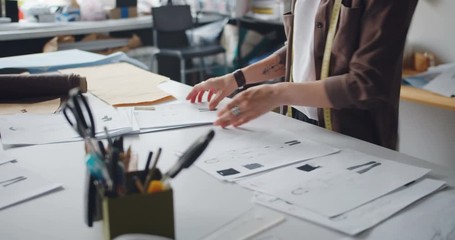 Clothes designer woman is holding drawings of clothes working in studio creating stylish garments. Cozy workshop with equipment and tools visible in background.