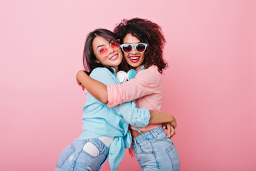 Excited slim girl with african hairstyle embracing asian female friend in trendy colorful...