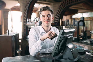 small business, people and service concept - happy man or waiter in apron at counter with cashbox working at bar or coffee shop.