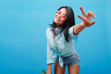 Gorgeous black-haired female model in vintage outfit posing with peace sign on blue background. Catching asian lady in sunglasses spending time on photoshoot and cute smiling.