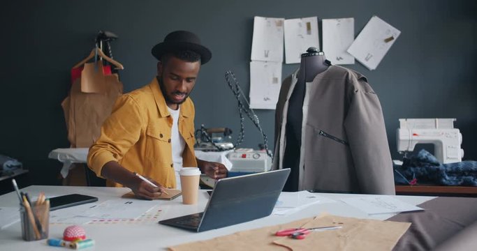 Trendy tailor African American man is measuring garment on dummy then writing in notebook working alone in workshop. Creative people, workplace and job concept.