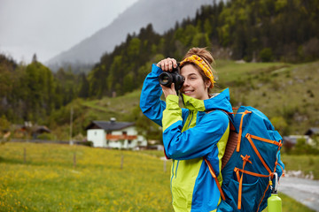 Outdoor shot of active young female tourists strolls in countryside, enjoys wonderful scenic view, mountain forest and houses in background, makes photo via camera. Adventurer explores nature
