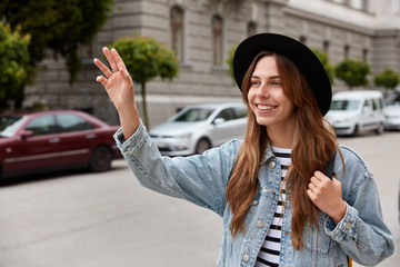 Young smiling young European woman walks outdoor, waves hand as notices friend into distance, strolls across street, wears stylish black headgear and denim jacket, poses over urban background.