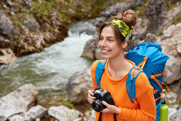 Adventurous female enjoys travel adventure, has hiking activity and nature tour, makes photo of landscapes, holds professional camera, carries rucksack, admires mountain brook, focused into distance