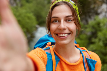 Tourist female hiker makes selfie portrait, smiles at camera, stands outdoor near nature outlook, wears casual comfortable clothes, being in good mood. Adventurious trip and spare time concept