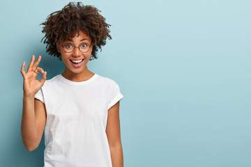 Horizontal shot of cheerful Afro American woman makes okay gesture, agrees with good proposal, wears spectacles and white casual t shirt, says incredible offer, isolated over blue background - obrazy, fototapety, plakaty