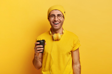 Handsome cheerful man uses headphones holds takeaway coffee, being in good mood, wears stylish yellow headgear and t shirt, has fun while enjoys aromatic drink, poses in studio. Monochrome shot