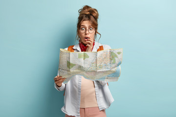 Half length shot of scared embarrassed woman looks stressfully at map, afraids of being lost, wears...