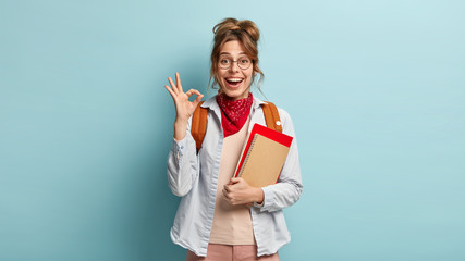 Indoor shot of delighted happy student makes okay gesture, demonstrates agreement, says everything is ok, holds textbooks or notepads, wears round optical spectacles and red bandana around neck