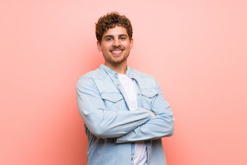 Blonde man over pink wall keeping the arms crossed in frontal position