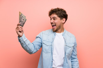 Blonde man over pink wall taking a lot of money