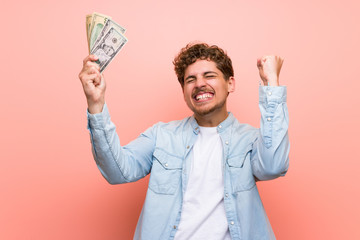 Blonde man over pink wall taking a lot of money