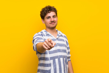 Blonde man over yellow wall points finger at you with a confident expression
