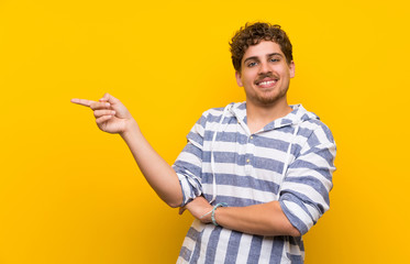 Blonde man over yellow wall pointing finger to the side in lateral position