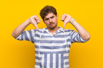 Blonde man over yellow wall showing thumb down with both hands