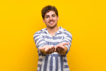 Blonde man over yellow wall holding copyspace imaginary on the palm to insert an ad