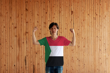 Man wearing Sudan flag color of shirt and standing with raised both fist on the wooden wall background.