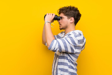 Blonde man over yellow wall and looking in the distance with binoculars