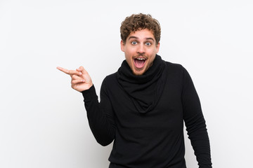 Blonde man over isolated white wall surprised and pointing finger to the side