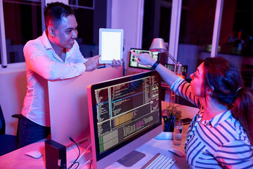 Smiling young businessman showing blank screen of digital tablet to his colleague while she sitting in front of computer and pointing at touchscreen in dark office.
