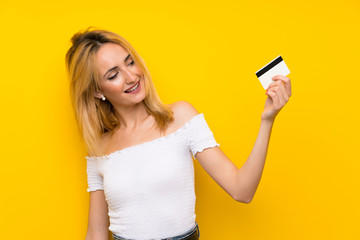 Naklejka premium Young blonde woman over isolated yellow wall holding a credit card