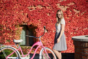 Fototapeta na wymiar Young pretty smiling blond woman in casual clothing and glasses posing at pink lady bicycle on bright warm sunny day in the fall on background of brick wall overgrown with beautiful red ivy leaves.