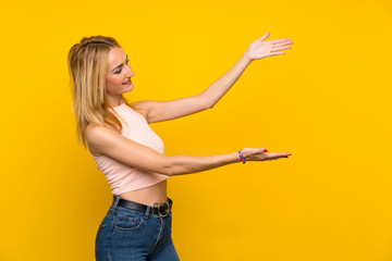 Young blonde woman over isolated yellow wall extending hands to the side for inviting to come