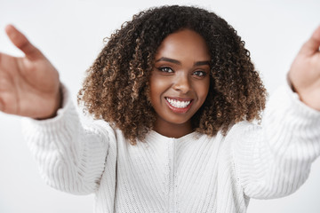 Girl wanna warm hugs. Tender and cute african-american female in sweater extending hands towards camera to cuddle and welcome friend smiling delighted with gentle gaze over white background - obrazy, fototapety, plakaty