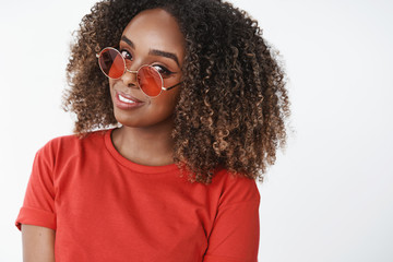 Close-up shot of flirty and sensual feminine good-looking african american woman in stylish sunglasses tilting head and gazing amused at camera with bright white smile, wearing red t-shirt