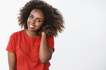 Waist-up shot of tender and romantic beautiful african-american woman with afro haircut holding hand behind on neck and smiling shy blushing flirty posing over white background in red t-shirt - obrazy, fototapety, plakaty
