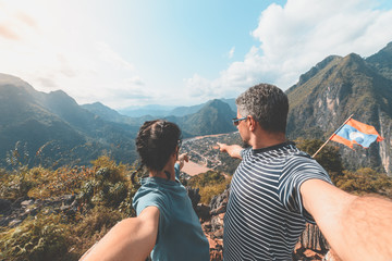 Naklejka premium Couple selfie on mountain top at Nong Khiaw panoramic view over Nam Ou River valley Laos travel destination in South East Asia, mature people traveling millenials concept, teal orange toned