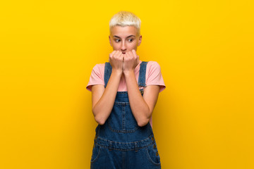 Teenager girl with overalls on yellow background nervous and scared putting hands to mouth