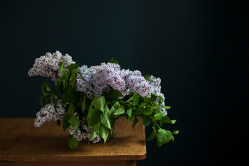 bouquet of lilac twigs in a transparent green jar on the wooden table opposite of black wall