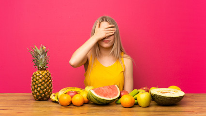 Young blonde woman with lots of fruits covering eyes by hands