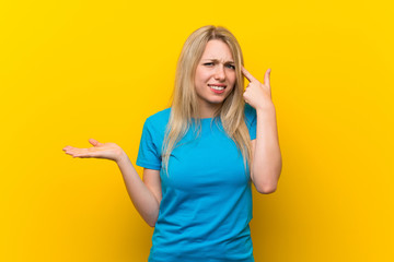 Young blonde woman over isolated yellow background making the gesture of madness putting finger on...