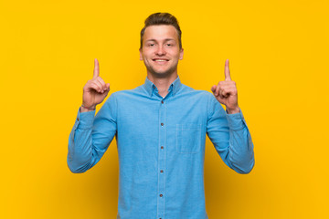 Blonde man over isolated yellow wall pointing up a great idea