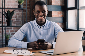 African guy working in cafe holding smartphone in hands and looking at camera with smile