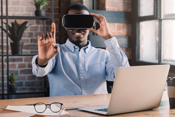 Young african business man wearing virtual reality headset, pointing in the air with finger while sitting at his desk in modern loft office