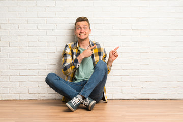 Blonde man sitting on the floor frightened and pointing to the side