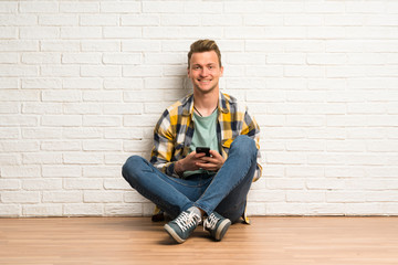 Blonde man sitting on the floor sending a message with the mobile