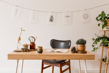 Fototapeta na wymiar Stylish scandinavian home interior of open space, with a lot of plants, design accessories, bamboo shelf, wooden desk and hanging mock up forest drawings . Botany concept of home decor. Sunny room.