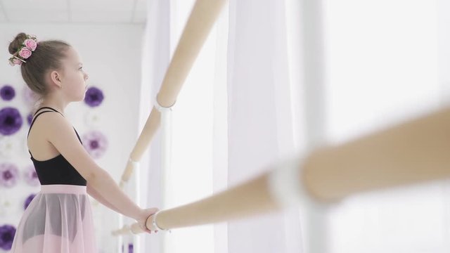 Dolly move of thoughtful little girl standing near ballet barre in the studio.