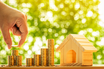 Woman hand hold a  stacked gold coin and wooden home model put on the table in the public park,...