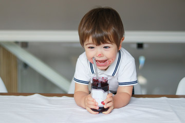 Portrait of funny happy little boy with smeared face eating cherry jam from glass jar with big spoon. Sweet tooth. Child nutrition concept.